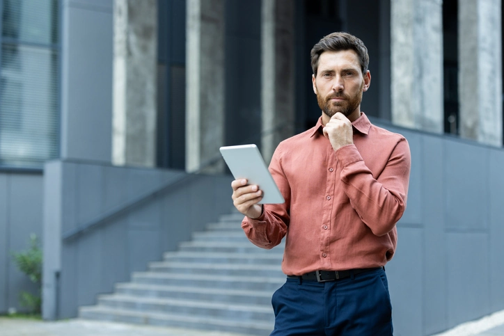 A businessman stands thoughtfully outside an office building with a digital tablet in hand